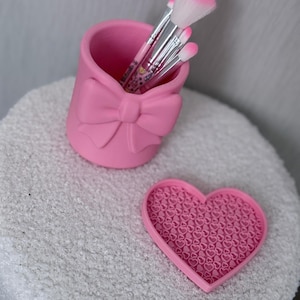 Puede incluir: Un portalápices rosa para brochas de maquillaje con un diseño de lazo, que contiene varios pinceles de maquillaje con cerdas rosas y blancas. Un limpiador de brochas rosa en forma de corazón con un patrón de corazones se encuentra junto al portalápices. Los artículos están sobre una superficie blanca y texturizada.