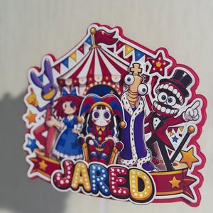 May include: A multi-layered paper cake topper featuring a circus tent background and five cartoon characters. The design includes a purple rabbit, a girl in a blue dress, a jester, a king, and a ringmaster. The name JARED is printed in large letters.