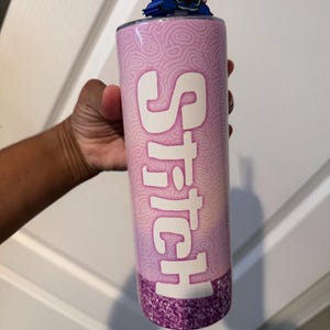 May include: A pink tumbler with the word "Stitch" in white, featuring a blue character figure on top. The bottom of the tumbler has a glittery purple design. A clear straw is visible.