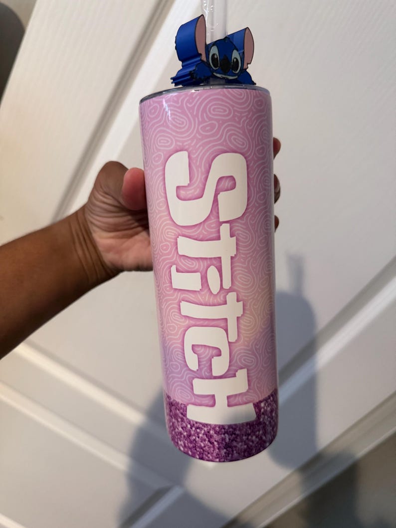 May include: A pink tumbler with the word "Stitch" in white, featuring a blue character figure on top. The bottom of the tumbler has a glittery purple design. A clear straw is visible.