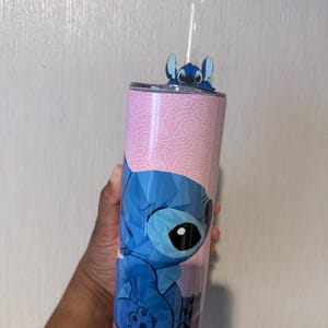 May include: A stainless steel tumbler featuring a blue geometric design of the character Stitch from Lilo & Stitch. The tumbler has a pink background with a swirl pattern and a glittery purple base. A small Stitch figure sits on the lid, and a clear straw is inserted.