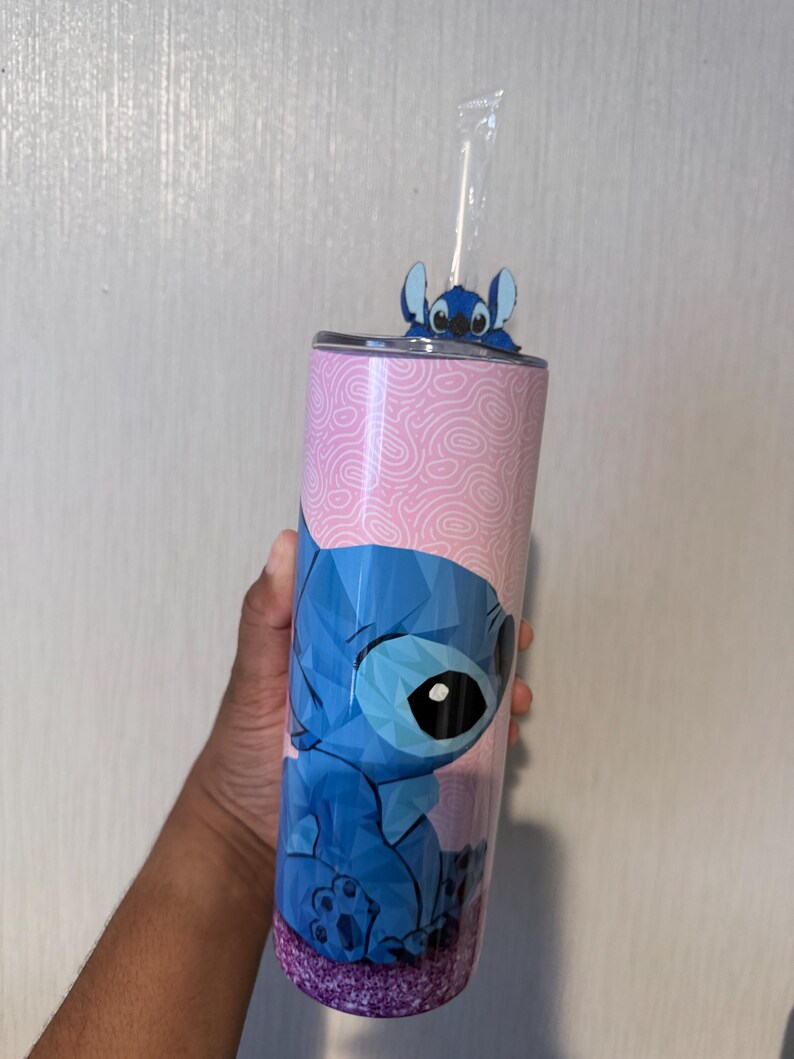 May include: A stainless steel tumbler featuring a blue geometric design of the character Stitch from Lilo & Stitch. The tumbler has a pink background with a swirl pattern and a glittery purple base. A small Stitch figure sits on the lid, and a clear straw is inserted.
