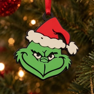 Puede incluir: Un adorno de personaje verde con una amplia sonrisa traviesa y ojos entrecerrados, que lleva un gorro de Papá Noel rojo con borde blanco. La decoración plana de goma cuelga de una cinta roja fina y tiene un contorno negro definido.