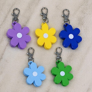 Puede incluir: Cinco llaveros con forma de flor en varios colores: morado, amarillo, azul, celeste y verde. Cada llavero tiene un centro blanco y un cierre plateado. Los llaveros están hechos de un material sólido.