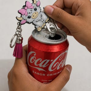 Puede incluir: Un abrelatas de metal plateado con un dije de vaca de dibujos animados con un lazo rosa y una borla rosa a juego. El abrelatas está unido a un llavero de metal y se muestra levantando la lengüeta de una lata roja de Coca-Cola de 350 ml.