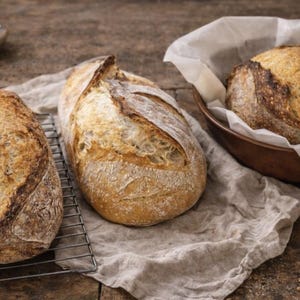 Può includere: Tre pagnotte di pane appena sfornate con crosta dorata. Una pagnotta poggia su una griglia di raffreddamento in metallo, un'altra su un panno di lino e la terza in una ciotola di legno foderata con carta pergamena. Una piccola ciotola di cereali e spighe di grano sullo sfondo.