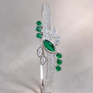 May include: Silver bracelet with a central design of a large, emerald-green gemstone and smaller green and clear gemstones. The bracelet band is encrusted with small, clear gemstones, creating a sparkling effect. The design is symmetrical and elegant.