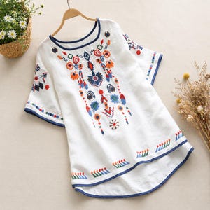 May include: White embroidered top with a round navy blue trimmed neckline and sleeves. The top is decorated with colorful floral embroidery in shades of orange, blue, red, and green. The top is hanging on a wooden hanger.