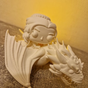 May include: A white figurine of a woman riding a dragon. The woman has her hair up and is wearing a dress. The dragon has large wings and a detailed head. The figurine is made of a solid, smooth material.