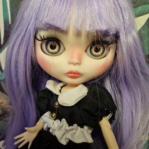 May include: A doll with long, lavender hair and large, expressive eyes. The doll wears a black dress with a white collar and gold buttons. The doll's face is painted with rosy cheeks and a delicate pout.