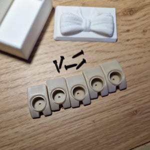 Könnte beinhalten: Eine Sammlung von 3D-gedruckten Elementen: eine weiße Box, ein Fliegen-Design, sechs beige Montagehalterungen und schwarze Schrauben. Die Artikel sind auf einer Holzoberfläche angeordnet, was auf ein DIY- oder Bastelprojekt hindeutet.