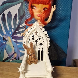 May include: A doll with red hair and a white dress is seated on a white decorative chair. A small brown bunny figurine sits on the chair. The background features a blue canvas with a parrot illustration.