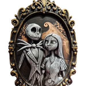 Könnte beinhalten: Eine dreidimensionale ovale Wandplakette mit einer skulpturalen Darstellung einer großen, dünnen Skelettfigur im Nadelstreifenanzug und einer weiblichen Figur mit langem Haar, beide in Grautönen, in einem verzierten, schwarz-goldenen Rahmen.
