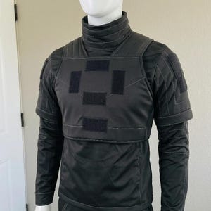 K&ouml;nnte beinhalten: Eine schwarze Weste im taktischen Stil &uuml;ber einem passenden lang&auml;rmeligen Shirt mit Stehkragen. Die Weste hat einen hohen Kragen, strukturierte Schulterpartien und vier rechteckige Klettverschl&uuml;sse auf der Brust. Details mit wei&szlig;en N&auml;hten.