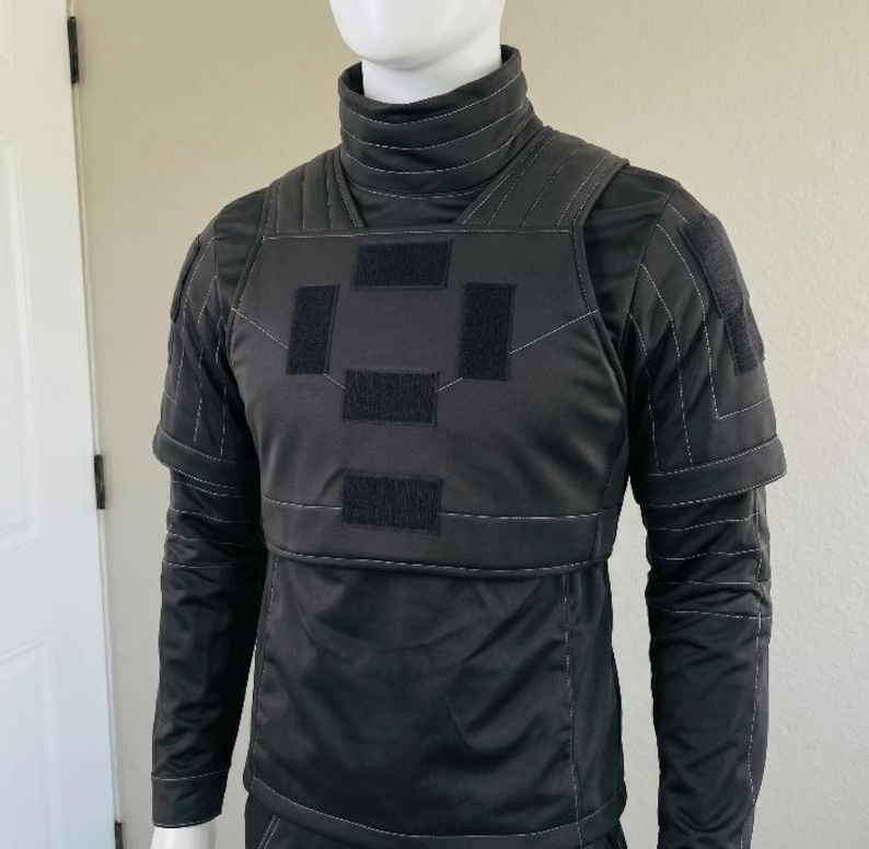 K&ouml;nnte beinhalten: Eine schwarze Weste im taktischen Stil &uuml;ber einem passenden lang&auml;rmeligen Shirt mit Stehkragen. Die Weste hat einen hohen Kragen, strukturierte Schulterpartien und vier rechteckige Klettverschl&uuml;sse auf der Brust. Details mit wei&szlig;en N&auml;hten.