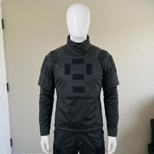 K&ouml;nnte beinhalten: Ein schwarzes taktisches Oberteil mit hohem Stehkragen, kurzen &Auml;rmeln mit verst&auml;rkten N&auml;hten und einem Brustbereich mit vier rechteckigen Klettverschluss-Patches in Kreuzform, pr&auml;sentiert auf einem wei&szlig;en Schaufensterpuppen-Torso.