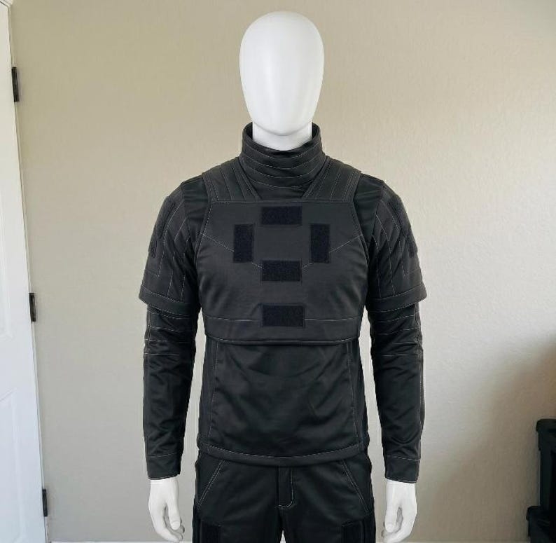 K&ouml;nnte beinhalten: Ein schwarzes taktisches Oberteil mit hohem Stehkragen, kurzen &Auml;rmeln mit verst&auml;rkten N&auml;hten und einem Brustbereich mit vier rechteckigen Klettverschluss-Patches in Kreuzform, pr&auml;sentiert auf einem wei&szlig;en Schaufensterpuppen-Torso.