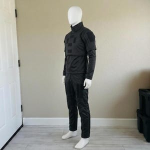 K&ouml;nnte beinhalten: Ein schwarzes taktisches Uniformset bestehend aus einem lang&auml;rmeligen Oberteil mit hohem Kragen und einer strukturierten Brustauflage mit rechteckigen Aufn&auml;hern sowie einer passenden schwarzen Hose mit verst&auml;rkten Eins&auml;tzen, pr&auml;sentiert auf einer wei&szlig;en Schaufensterpuppe.