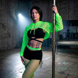 Könnte beinhalten: Eine Frau in einem neongrünen Mesh-Crop-Hoodie, einem schwarzen Sport-BH und schwarzen Shorts mit Kordelzugdetails. Sie trägt auch neongrüne Netzstrümpfe. Das Outfit ist für Tanz- oder Fitnessaktivitäten konzipiert. Sie hält eine Metallstange.