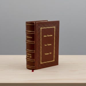May include: A brown leather-bound book titled "Anna Karenina" by Leo Tolstoy, Volume I-II. The book has gold lettering and decorative gold trim. A red ribbon bookmark is visible.