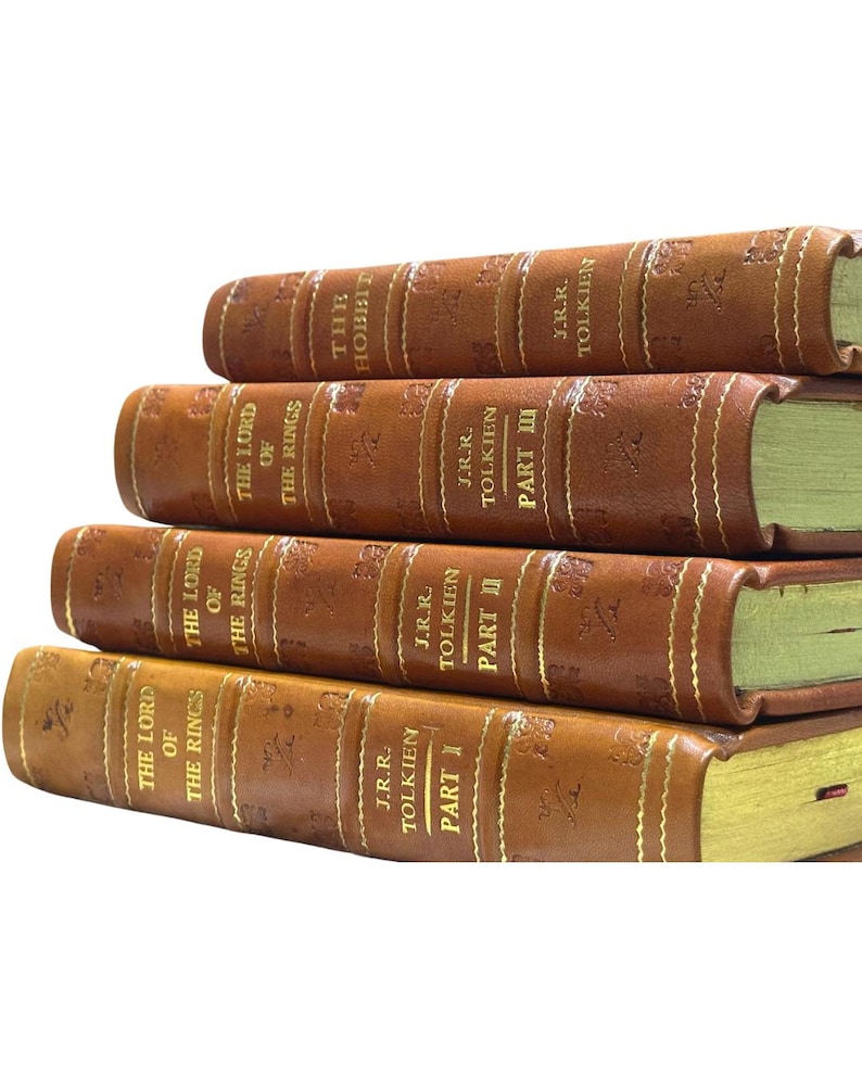 May include: A stack of leather-bound books with gold lettering. The titles include "The Hobbit" and "The Lord of the Rings" by J.R.R. Tolkien. The books are brown with gold-colored page edges.