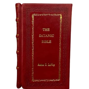 May include: A red hardcover book titled "The Satanic Bible" by Anton S. LaVey. The book has gold lettering and decorative gold trim around the title on the cover. The spine of the book is also red with gold accents.