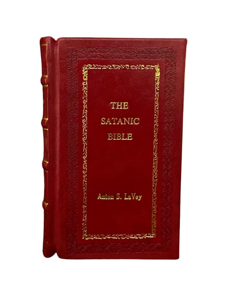 May include: A red hardcover book titled "The Satanic Bible" by Anton S. LaVey. The book has gold lettering and decorative gold trim around the title on the cover. The spine of the book is also red with gold accents.