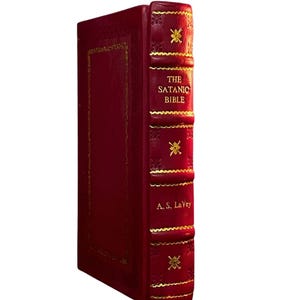 May include: A red hardcover book titled "The Satanic Bible" by A.S. LaVey. The book has gold accents and decorative elements on the spine and cover. The book is set against a white background.