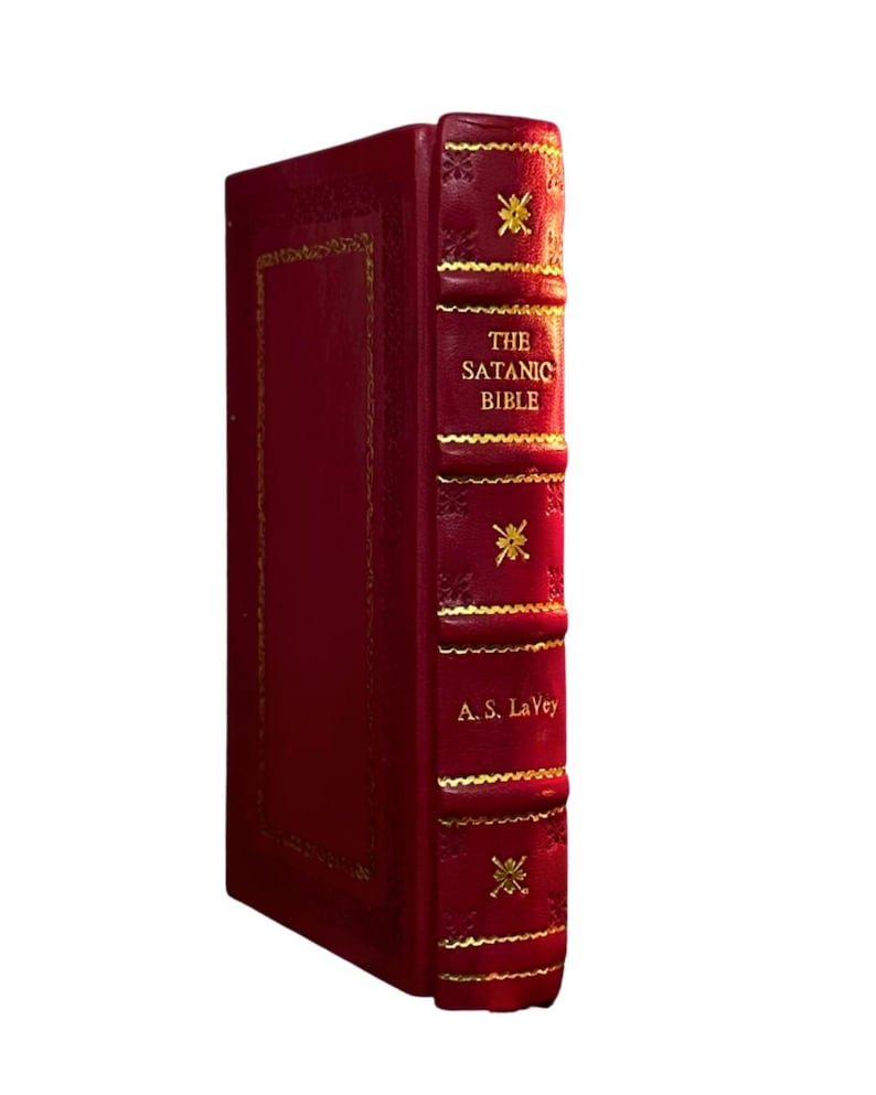 May include: A red hardcover book titled "The Satanic Bible" by A.S. LaVey. The book has gold accents and decorative elements on the spine and cover. The book is set against a white background.