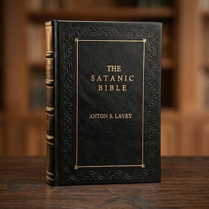 Puede incluir: Un libro de tapa dura negro titulado "The Satanic Bible" de Anton S. LaVey. El libro tiene letras doradas y un borde dorado en la portada. El lomo también es dorado. El libro está sobre una superficie de madera.