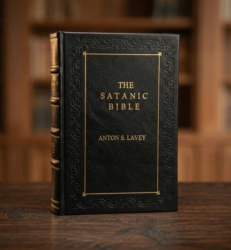 May include: A hardcover book titled "The Satanic Bible" by Anton S. LaVey. The book has a black cover with gold lettering and a gold border. The spine is also gold. The book is on a wooden surface.