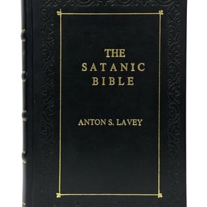 May include: A hardcover book titled "The Satanic Bible" by Anton S. LaVey. The book has a black cover with gold lettering and a decorative gold border. The spine is also black.