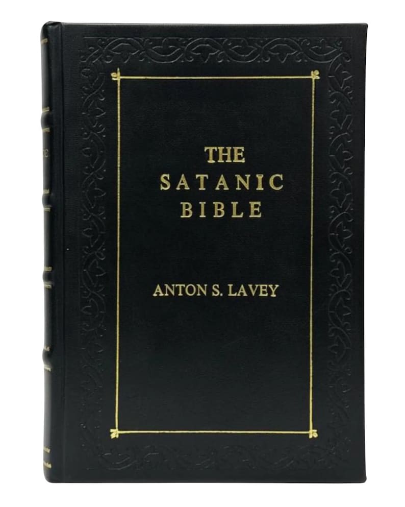 May include: A hardcover book titled "The Satanic Bible" by Anton S. LaVey. The book has a black cover with gold lettering and a decorative gold border. The spine is also black.