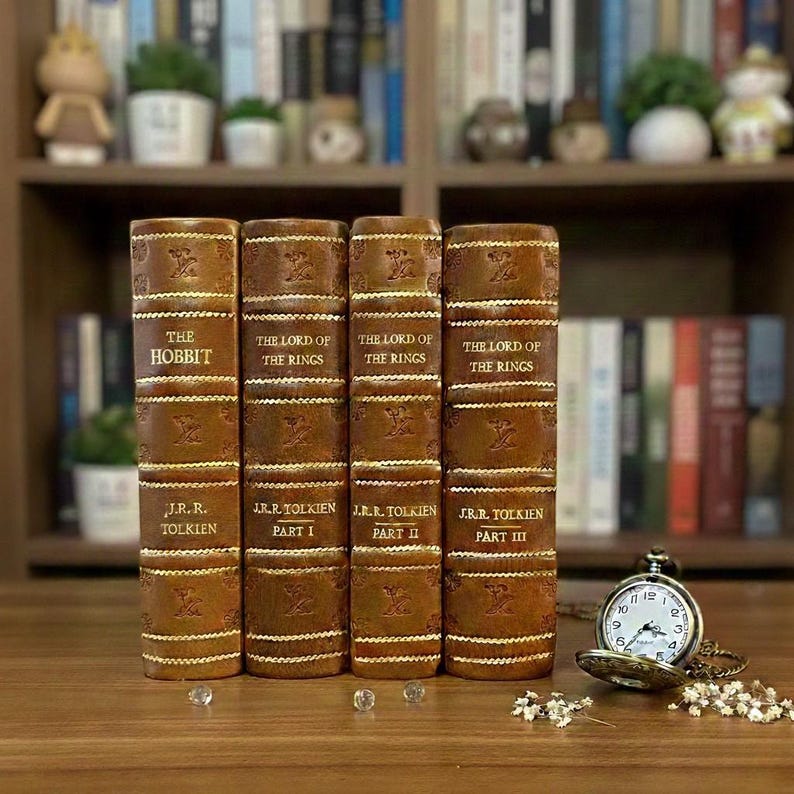 May include: Four leather-bound books, including "The Hobbit" and "The Lord of the Rings" trilogy, are displayed upright. The books are brown with gold lettering and decorative details. A pocket watch and small crystals are on the wooden surface in front of the books.