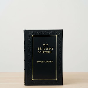 Op de afbeelding: Een zwart hardcover boek met de titel "The 48 Laws of Power" van Robert Greene. Het boek heeft gouden letters en een gouden rand. Het boek ligt op een lichtbruin houten oppervlak.