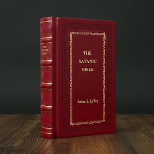 May include: A hardcover book titled "The Satanic Bible" by Anton S. LaVey. The book has a red cover with gold lettering and decorative gold trim. The spine also features gold lettering. The book is displayed upright.