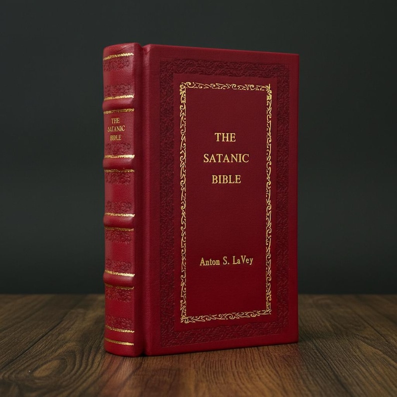 May include: A hardcover book titled "The Satanic Bible" by Anton S. LaVey. The book has a red cover with gold lettering and decorative gold trim. The spine also features gold lettering. The book is displayed upright.