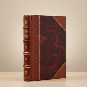 May include: A hardback book with a brown leather cover and gold detailing. The spine displays the title "The Religion of The Egnor Name Aertles Romaines". The cover features a burgundy marbled pattern.