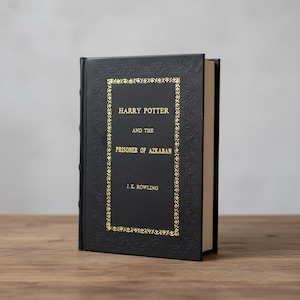 Può includere: Un libro con copertina rigida nera intitolato "Harry Potter e il Prigioniero di Azkaban" di J.K. Rowling. Il titolo e il bordo sono dorati. Il libro è appoggiato su una superficie di legno.