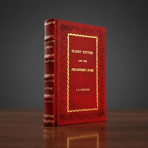 May include: A hardcover book titled "Harry Potter and the Philosopher's Stone" by J.K. Rowling. The book has a red cover with gold lettering and decorative borders. The spine is also red with gold accents and the author's name.