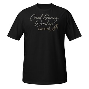 Puede incluir: Camiseta negra con la frase "Cried During Worship (AGAIN)" en letra cursiva, adornada con un diseño floral. La camiseta tiene cuello redondo y mangas cortas.