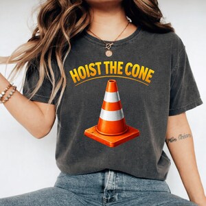 May include: Dark grey t-shirt with a graphic of an orange and white traffic cone on a square base. The text "HOIST THE CONE" is arched above the cone in yellow letters.