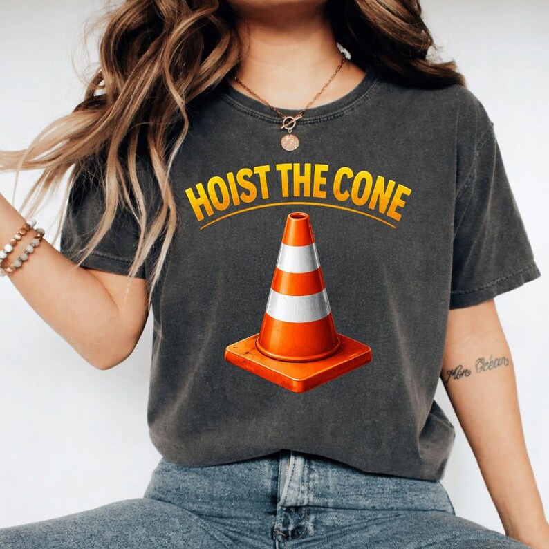 May include: Dark grey t-shirt with a graphic of an orange and white traffic cone on a square base. The text "HOIST THE CONE" is arched above the cone in yellow letters.