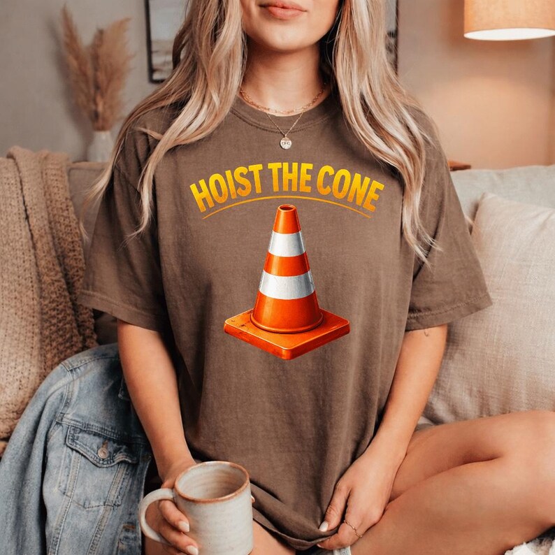 May include: Brown t-shirt featuring the text "HOIST THE CONE" in yellow above a graphic of an orange and white traffic cone. The cone is on a square orange base. The shirt is a casual style.