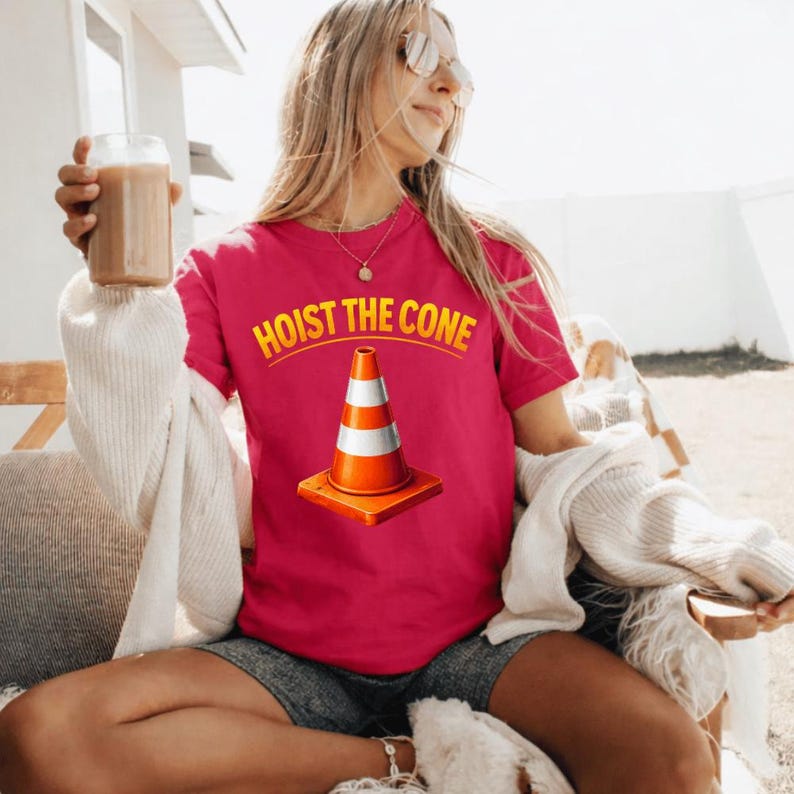 May include: A magenta t-shirt featuring the text "HOIST THE CONE" in yellow above an orange and white traffic cone graphic. The shirt is made of a soft material.
