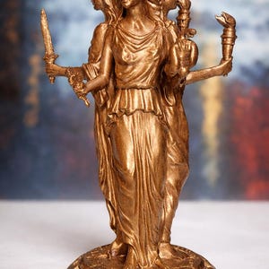 Può includere: Statua color bronzo raffigurante una figura a tre facce, ognuna con un'espressione diversa. La figura centrale tiene una spada, mentre le altre tengono una torcia e un vaso. La statua è su una base circolare.