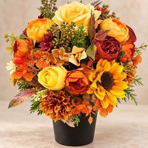 Könnte beinhalten: Ein Blumenarrangement in einem schwarzen Topf mit gelben Rosen, orangefarbenen Pfingstrosen, einer Sonnenblume und dunkelroten Blüten. Das Bouquet enthält grünes Blattwerk, kleine Beeren und herbstlich gefärbte Blätter in einer dichten Form.