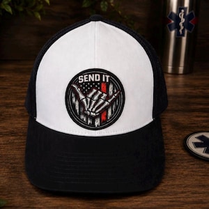 Op de afbeelding: Zwarte en witte truckerpet met een wit voorpaneel en een zwarte mesh achterkant. De pet heeft een cirkelvormige patch met de tekst "SEND IT" boven een skelet handgebaar, met rode en witte strepen.
