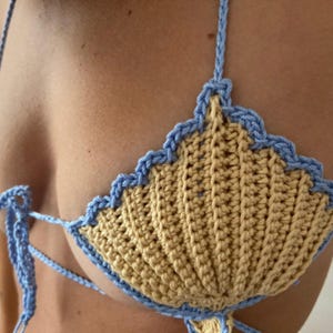 May include: A handmade crochet bikini top in a seashell design. The top features a light yellow base with a scalloped edge and straps in light blue. The crochet work is visible, showcasing the texture and craftsmanship of the garment.