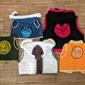 Puede incluir: Colección de cinco chalecos de ganchillo hechos a mano en varios colores. Cada chaleco presenta un símbolo diferente, incluyendo agua, tierra, fuego y aire. Los chalecos son en tonos de azul, negro, verde, blanco y naranja.